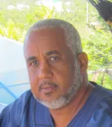 A Black man with greying black hair and beard looks into the camera with greenery behind him. He is wearing a blue shirt.