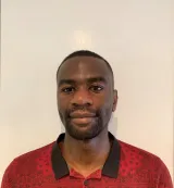 A Black person with very short black hair looks into the camera with a slight smile. They are wearing a red shirt with a printed black line drawing design and a black collar.