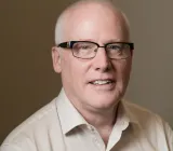 A middle-aged man with short grey hair and glasses, wearing a white shirt and smiling warmly at the camera.