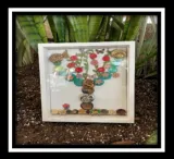 An art design in a square-shaped shadow box shows a foliage pattern with buttons in the shape of a tree. The art piece sits in soil with plants in the background.