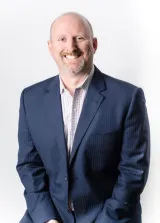 A white man with short hair and a goatee smiles towards the camera. He is wearing a dark blue suite and white checkered dress shirt.