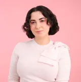 A white woman with brown hair smiles towards the camera. She is wearing a light pink sweater with a pink bow over her left shoulder.