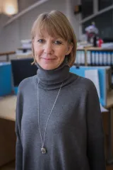 A white woman with blonde hair smiles towards the camera. She is wearing a long gray turtleneck sweater and a silver necklace and pendant.