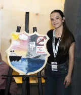 A young woman stands next to a shield painted with sunny skies and a boat floating on water on one side, and a dark sky and a sinking boat.