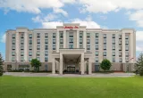 A large, modern hotel building with a light-coloured exterior and multiple rows of windows. The entrance features a covered portico supported by columns, and the red "Hampton Inn" sign is displayed at the top of the building. A green lawn stretches across the foreground under a partly cloudy sky.