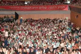 A packed auditorium filled with hundreds of attendees, many wearing teal scarves and badges, gathered beneath a large red banner reading 'The time has arrived: Peace is the only option' in Hebrew.