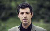 Headshot of a middle-aged man with dark curly hair wearing a grey blazer over a black shirt, photographed outdoors against a blurred green foliage background. His expression is serious and direct.