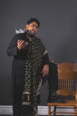 A bearded drag king in a black outfit with long black and gold-patterned scarf holds his right hand up with a jewelled crown on his shoulder, posing confidently in front of a grey wall beside a wooden chair.