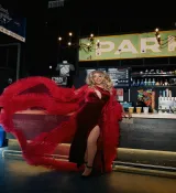 Une artiste drag aux longs cheveux blonds prend une pose théâtrale dans une robe rouge fluide qui ondule largement autour d’elle. Derrière elle, on aperçoit en arrière-plan un bar éclairé avec une enseigne partielle de l’établissement.