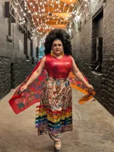 A boldly styled drag queen walks confidently down a narrow alley decorated with string lights overhead, wearing a red top, a colourful floral ribbon skirt, large black bouffant hair, yellow earrings, and a flowing shawl spread wide on each side