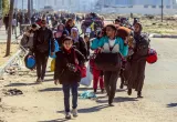Families carrying bags walk along a dirt road.