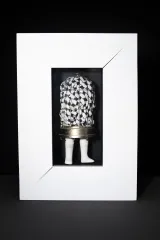 A mixed-media sculpture consisting of a round-top metal birdcage with white doll’'s legs attached to the base. The cage encloses a white cloth with black patterns. The entire assemblage is presented in a white frame.