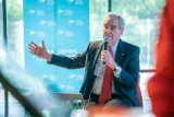 Man in a suit and red tie speaks into a microphone and gestures, seated before a Central European University backdrop.