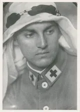 White man in an army uniform with a black cross at the edge of his collar wears a keffiyeh, white headdress worn in the Middle East.