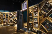 A large, free-standing art installation contains hundreds of objects. It is framed in cedar and there is an open door in the middle that people can walk through.