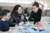 A smiling parent and two children sit at a table, with crafting supplies including markers, scissors, glue and construction paper laid out before them.