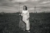 A woman in a long white dress stands in a field holding a bandura, a Ukrainian plucked string folk instrument.