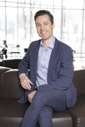 A caucasian man in a business suit has dark hair and blue eyes. He is sitting cross-legged.