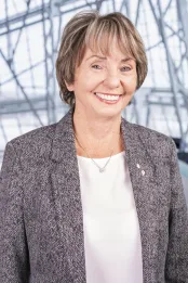 A white woman with light brown hair is wearing a grey patterned blazer and a white shirt. She is smiling facing the camera and standing in a relaxed manner.