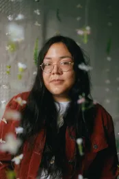 Veronica Blackhawk, a young Anishinaabe person with long dark hair and round glasses, is wearing a red corduroy jacket.