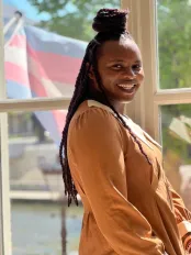 A Black woman stands smiling in front of a window with the trans flag of Pride in the background. Her long black hair is braided and she is wearing a sandstone orange garment.