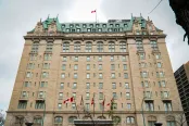 A large historic hotel building with a beige stone façade, green copper roof, and ornate architectural details. A Canadian flag is flying at the top of the building and several flags are displayed at the entrance below.
