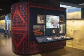A Museum exhibit wall with several artifacts displayed behind glass and a video being shown in the middle. The wallpaper on the display features a navy and orange Palestinian embroidery style called tatreez. 