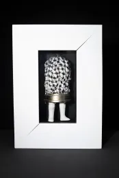 A mixed-media sculpture consisting of a round-top metal birdcage with white doll’'s legs attached to the base. The cage encloses a white cloth with black patterns. The entire assemblage is presented in a white frame.
