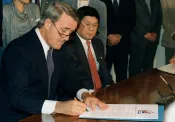 Brian Mulroney sitting and signing a document. Art Miki is sitting beside him looking on.