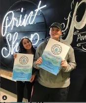 A young woman and man hold up posters that promote a fashion show. In the background are bright neon lights.
