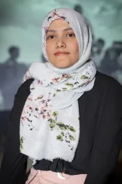 A portrait of a young woman with dark eyes wearing a black long-sleeved top. Her hair is covered with a mostly-white head scarf that has a green and pink floral pattern on it.