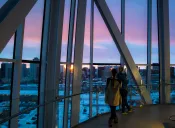Two people gaze out a tall glass window at a sunset over a cityscape.
