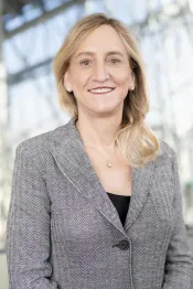 A white woman with light brown hair, light brown eyes and a white top smiles towards the camera.