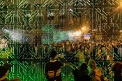 Five people stand in front of intricately patterned joined mirrors that reflect green lights and a throng of onlookers.