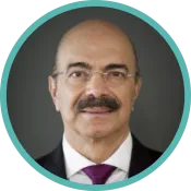 A man with a dark moustache and small, frameless glasses wears a dark suit and a purple tie.