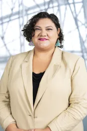 An Indigenous woman with short brown hair wears a beige blazer, layered over a black shirt. She is wearing pink-green-white beaded earrings. She is forward facing, and she is smiling at the camera.