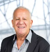 A white man with short white hair wears a black blazer, layered over a patterned dress shirt. He is facing forward and is smiling at the camera.