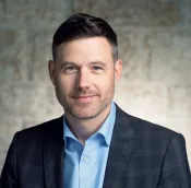 A white man in a checked suit jacket and blue shirt is posing for a professional headshot against a stone background. He has short dark hair and a trimmed beard.