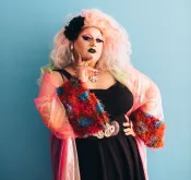 A curvy drag performer with voluminous pink hair, black eye and lip makeup poses with a hand on her hip. She wears a filmy pink jacket with colourful fur cuffs over a black dress.