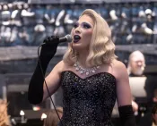An elegant blonde drag performer in a black sequined gown, diamond necklace and long black evening gloves sings into a microphone. 