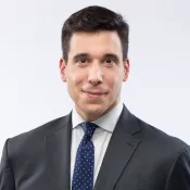 A white man with dark hair smiles slightly into the camera. He is wearing a dark suit with an off-white shirt and a dark blue tie with small white polka dots.