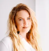 A white woman with long blond hair and a nose ring looks into the camera. She is wearing a white shirt.