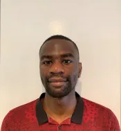 A Black person with very short black hair looks into the camera with a slight smile. They are wearing a red shirt with a printed black line drawing design and a black collar.