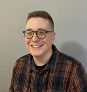 A white person with short dark hair and glasses smiles into the camera. They are wearing a dark green, dark yellow and orange tartan collared shirt.