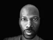 A black-and-white close-up portrait of a Black man looking straight at the camera with a neutral expression.