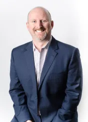 A white man with short hair and a goatee smiles towards the camera. He is wearing a dark blue suite and white checkered dress shirt.