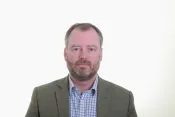 A white man with short greying hair and beard looking impartially at the camera. He is wearing a gray blazer with a blue and white checkered dress shirt.