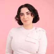 A white woman with brown hair smiles towards the camera. She is wearing a light pink sweater with a pink bow over her left shoulder.