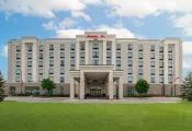 A large, modern hotel building with a light-coloured exterior and multiple rows of windows. The entrance features a covered portico supported by columns, and the red "Hampton Inn" sign is displayed at the top of the building. A green lawn stretches across the foreground under a partly cloudy sky.