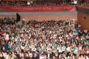 A packed auditorium filled with hundreds of attendees, many wearing teal scarves and badges, gathered beneath a large red banner reading 'The time has arrived: Peace is the only option' in Hebrew.